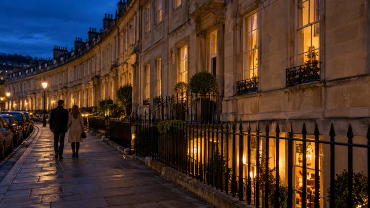 A curved Georgian terrace of honey-coloured Bath stone at night, with warm light from a basement bar below the railings