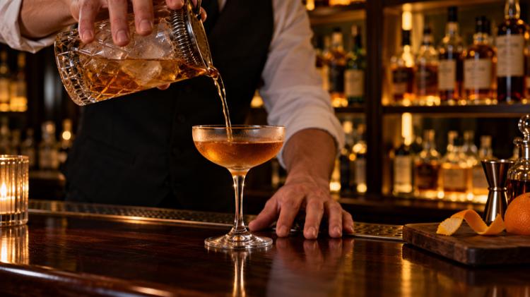 A bartender pouring a stirred amber cocktail into a coupe on a dark wooden bar, with whisky bottles lit behind
