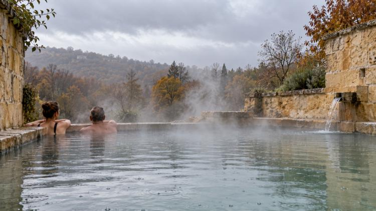Steam rising from a warm open-air stone pool in light rain, with two bathers at the edge and autumn hills behind