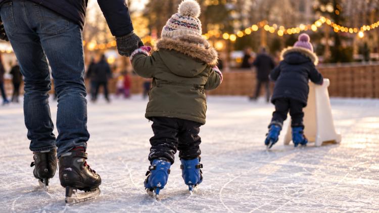 A small child in a bobble hat holding a parent's hand on an outdoor ice rink, with another child pushing a skate aid and fairy lights behind