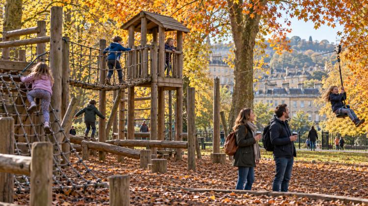 Children on a wooden adventure playground and zip line in a Bath park in autumn, with Bath stone houses beyond the trees