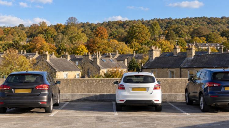 Cars parked on the open top deck of a multi-storey car park, with slate roofs and honey-coloured stone houses below a wooded hillside in Bath