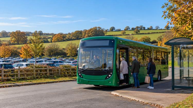 Shoppers boarding a green single-decker bus at a Park and Ride shelter, with rows of parked cars and green fields behind, outside Bath